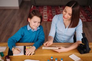 Aunt and nephew do homework together. Women explain kid writes in notebook. Schoolboy studying at home. Family relationships, primary education, childhood