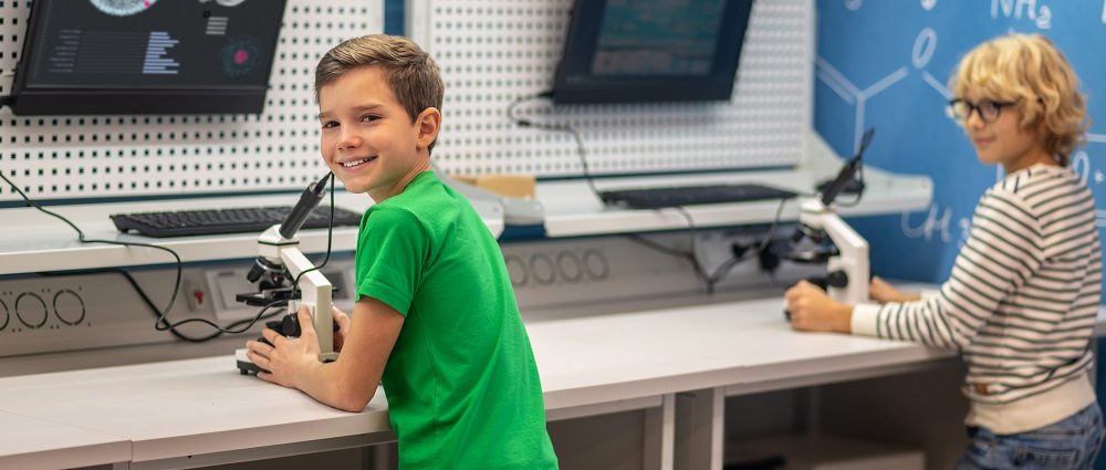 Joyful boy turned to camera standing near charge board with microscope and classmate studying in chemistry class
