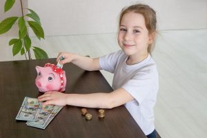little cute girl sits at a table on which banknotes and coins are laid out and puts money in a piggy bank, financial literacy.