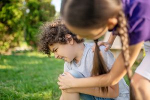 Dark-haired girl leaning over a young Caucasian boy suffering from the knee pain after the fall-down