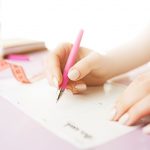 Female hands holding pen, writing. Side view on woman on trendy color pink desk. Woman and stilish workplace. Women's Day concept