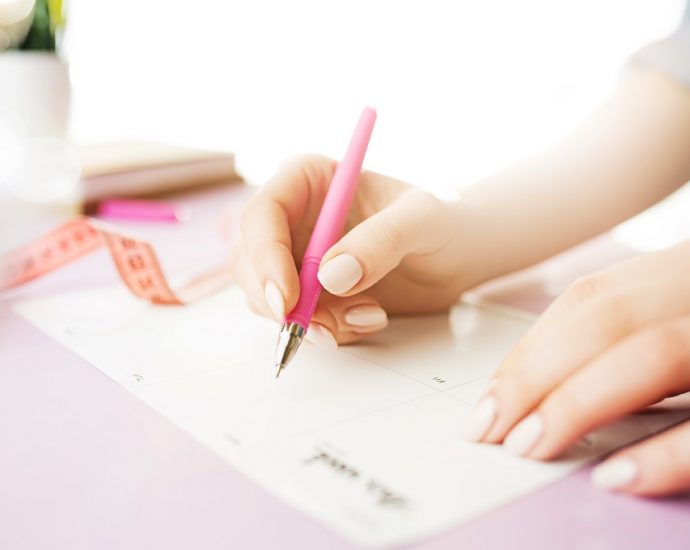 Female hands holding pen, writing. Side view on woman on trendy color pink desk. Woman and stilish workplace. Women's Day concept