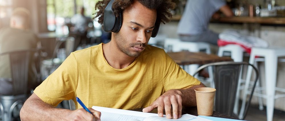Hipster Afro American guy doing his home assignment looking attentively in book and copybook listening to music in headphones and drinking coffee while sitting at cozy restaurant. Education concept