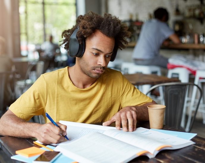 Hipster Afro American guy doing his home assignment looking attentively in book and copybook listening to music in headphones and drinking coffee while sitting at cozy restaurant. Education concept