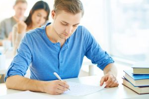 Portrait of handsome student carrying out test at lesson with groupmates on background