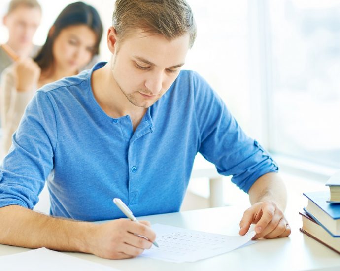 Portrait of handsome student carrying out test at lesson with groupmates on background