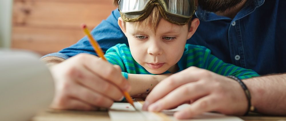 Boy in pilot eyewear looking at sketch of toy plane being drawn by his father