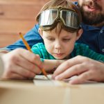 Boy in pilot eyewear looking at sketch of toy plane being drawn by his father
