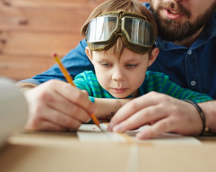 Boy in pilot eyewear looking at sketch of toy plane being drawn by his father