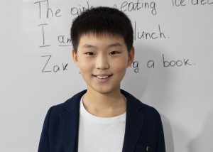 An Asian kid in front of a whiteboard with English sentences