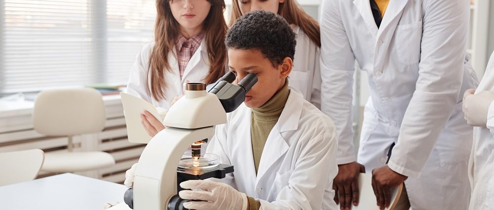 Portrait of young black girl looking into microscope with while doing experiments with group of children in school chemistry lab