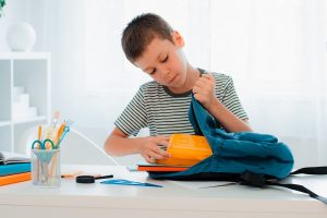 schoolboy putting school stationery into backpack at table indoors at home white room. preparation for school, homework