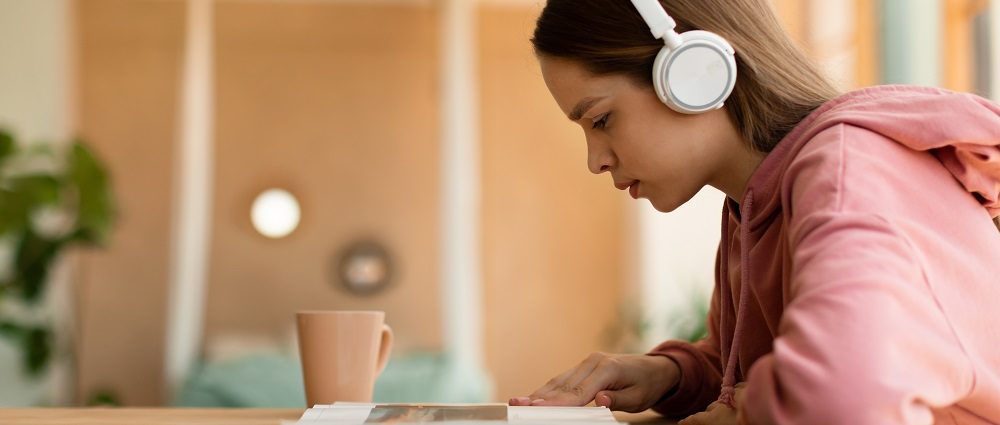 Smart focused teenager girl listening music in wireless headphones and reading book, sitting at desk in bedroom interior at home, side view, copy space